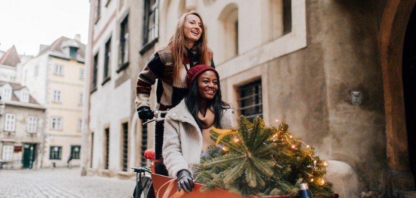 Zwei Freundinnen sind zusammen mit einem Lastenrad durch die Straßen unterwegs. Eine Person fährt das Rad, die andere Person sitzt drin. Sie transportieren einen kleinen Nadelbaum.