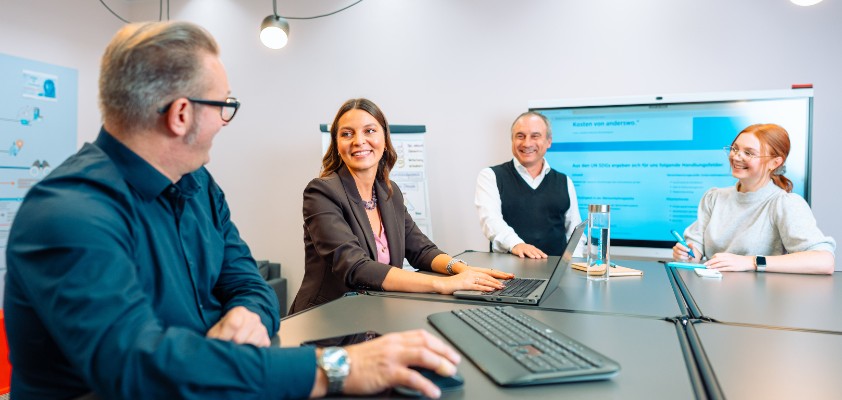 Mehrere Kolleginnen und Kollegen in einem Besprechungsraum schauen gemeinsam auf einen Laptop 