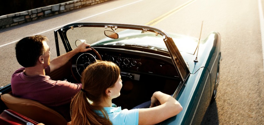 Eine Frau und ein Mann fahren in einem blauen Oldtimer-Cabrio durch den Sonnenschein.