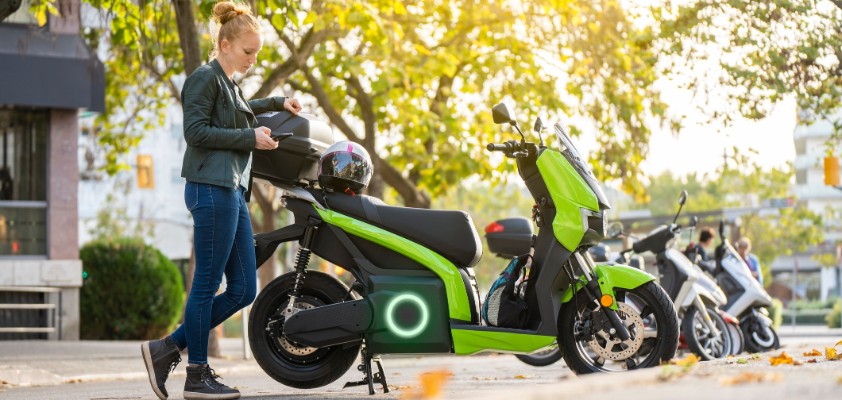 Eine blonde Frau steht neben ihrem elektrischen Motorrad und hält ihren Helm in der Hand. 