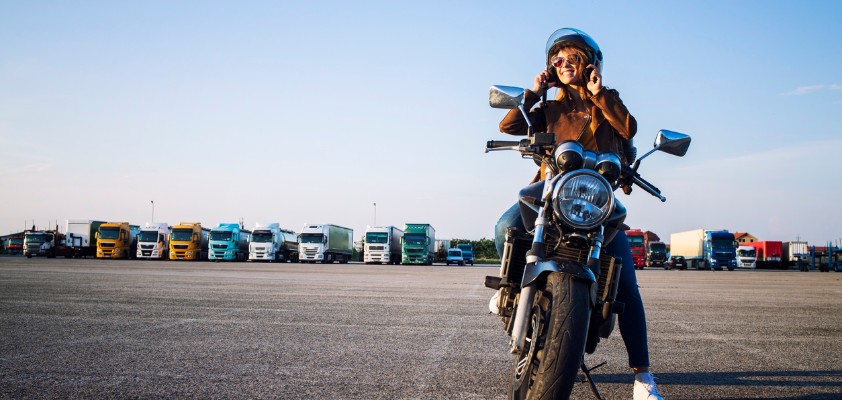 Frau sitzt mit Helm auf einem Motorrad auf einem großen Parkplatz, im Hintergrund stehen Lkw.
