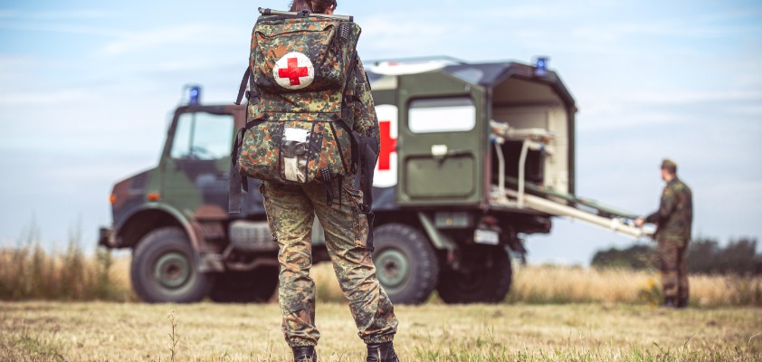 Eine Soldatin mit Sanitätsrucksack schaut in die Richtung eines Sanitätsfahrzeugs des Militärs