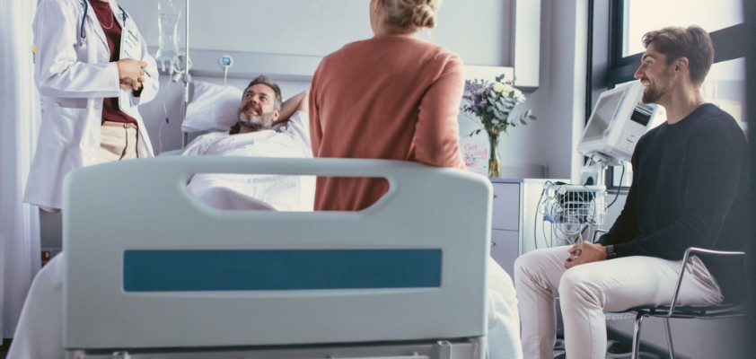 Ein männlicher Patient liegt in einem Krankenhausbett. Rechts von ihm steht eine weibliche Ärztin, eine Frau sitzt am Fußende des Bettes und ein Mann daneben auf einem Stuhl. 