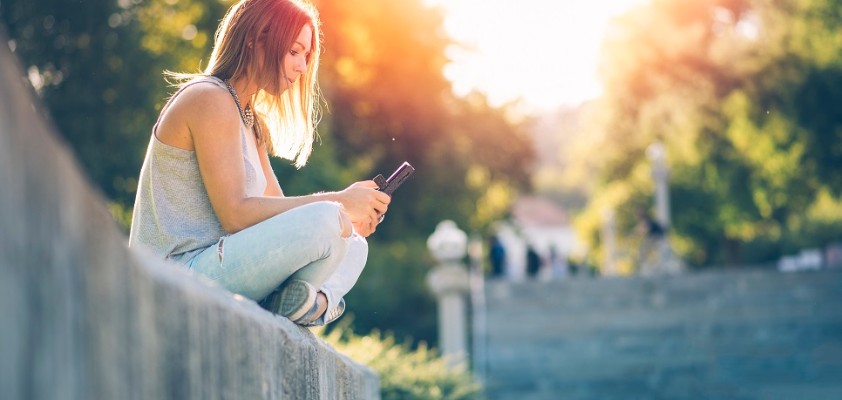 Ein Mädchen sitzt auf einer Steinmauer und schaut auf ihr Mobiltelefon. 