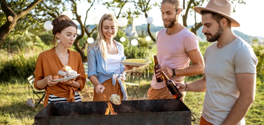 Eine Gruppe von Freunden steht zu viert an einem Holzgrill im Grünen. 