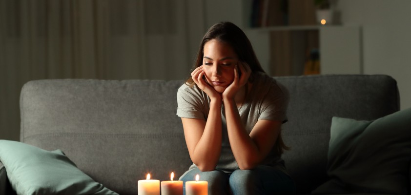 Eine junge Frau sitzt in ihrem Wohnzimmer ohne Strom und schaut ernüchternd auf drei leuchtende Kerzen auf dem Wohnzimmertisch. 