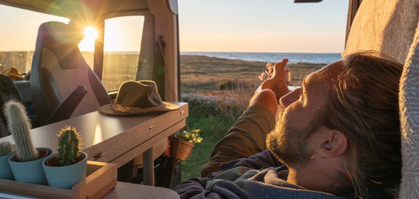 Ein junger Mann liegt in seinem ausgebauten Campervan mit Sonne im Hintergrund.
