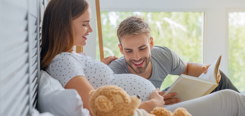 Eine schwangere Frau liegt im Bett. Ein Mann liegt neben ihr und liest ein Bett vor, während er auf den Babybauch schaut. 