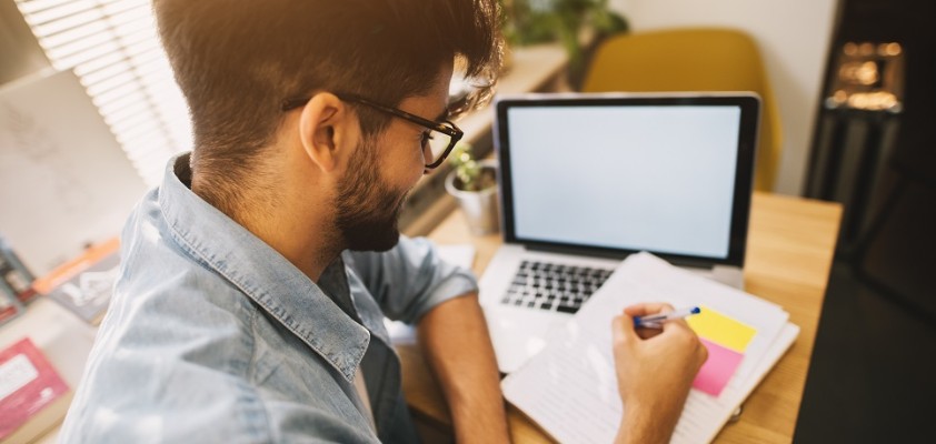 Ein junger Mann sitzt mit Laptop am Schreibtisch und liest sich ein ausgedrucktes Dokument durch. 