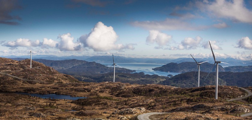 Landschaft mit Windkraftanlagen auf Hügeln und Blick auf das Meer im Hintergrund.
