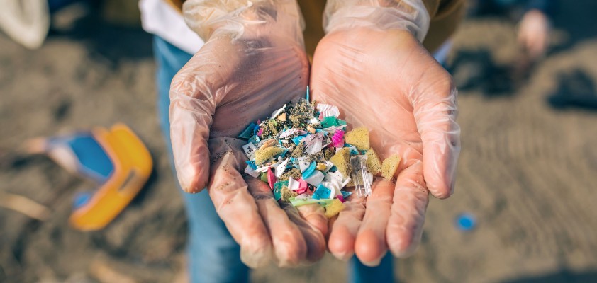 Zwei Hände halten kleine Teile aus Mikroplastik. Im Hintergrund ist ein Strand zu erkennen. 