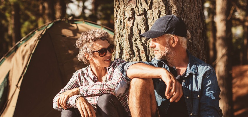 Eine Frau ein Mann in Wanderkleidung sitzen nebeneinander angelehnt an einen Baum und unterhalten sich.