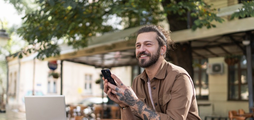 Ein Mann mit Tattoos am Arm sitzt im Außenbereich eines Cafés, hat eine Tasse vor sich stehen und blickt lachend Richtung Straße.