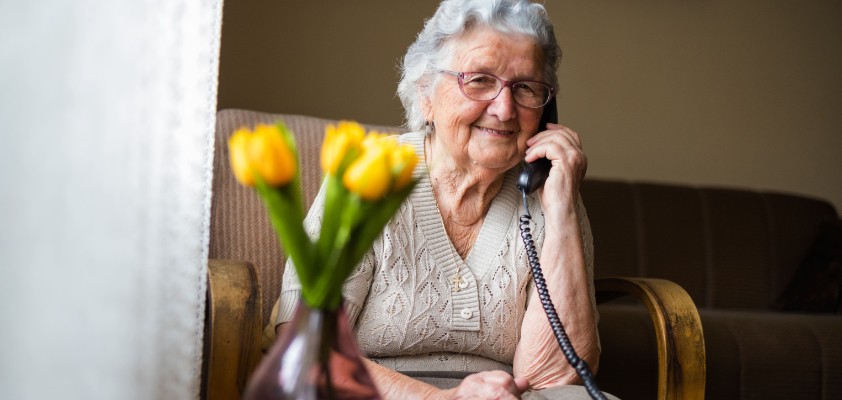 Eine ältere Dame sitzt in ihrem Zuhause am Kabeltelefon. 