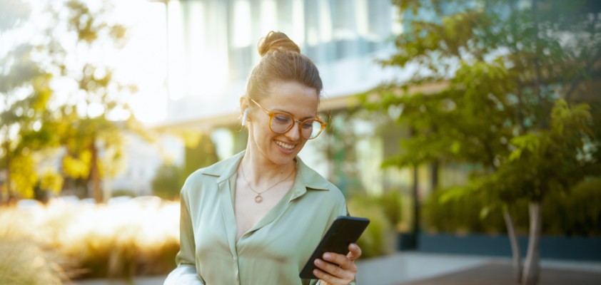 Eine junge Frau verlässt Bürogebäude und schaut glücklich auf ihr Telefon.
