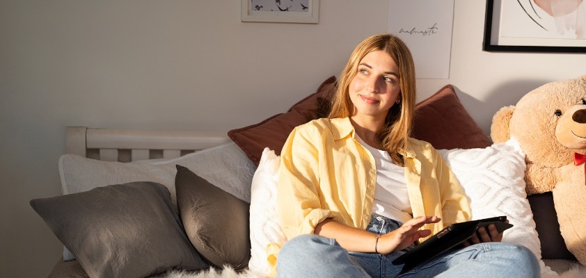 Eine junge Frau sitzt mit einem Riesen-Teddy auf ihrem Bett und hält ein Tablet in der Hand.