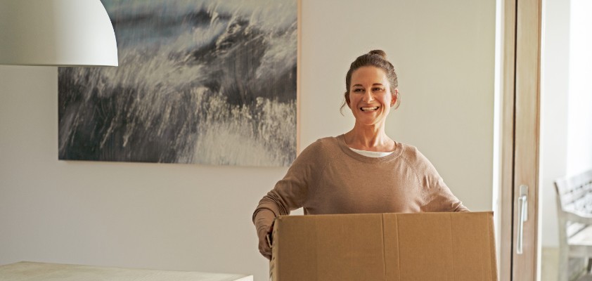 Eine Frau trägt lächelnd einen Umzugskarton durch ein Wohnzimmer.
