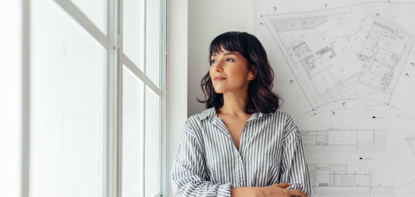 Eine junge Frau lehnt an einer Wand, an der Grundrisse hängen, während sie nach rechts aus dem Fenster schaut.