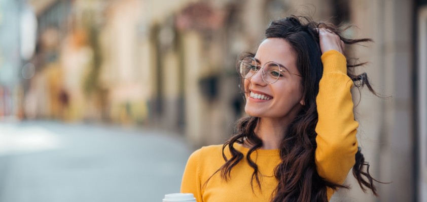 Eine junge erwachsene Frau steht in einer Straßengasse und hält in ihrer rechten Hand ein Heißgetränk im Pappbecher.
