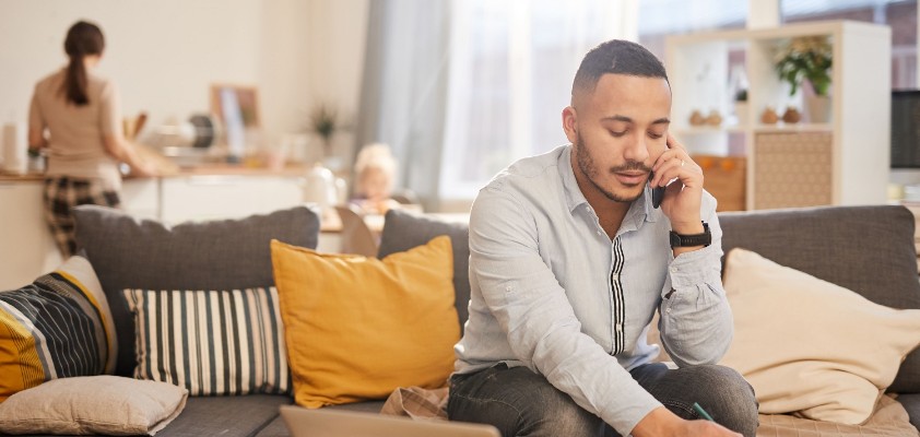 Ein junger Mann sitzt mit seinem Laptop am Wohnzimmertisch und telefoniert, während er sich handschriftlich Notizen macht. 
