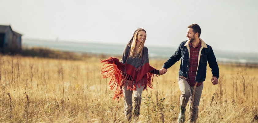 Eine junge Frau und ein junger Mann gehen lächelnd Hand in Hand über eine Wiese.