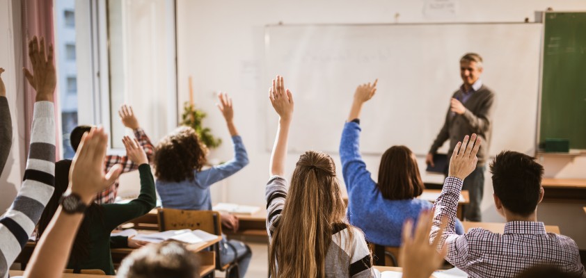 Ein Lehrer steht vor eine Klasse in einem Klassenzimmer. Viele Schüler melden sich. 
