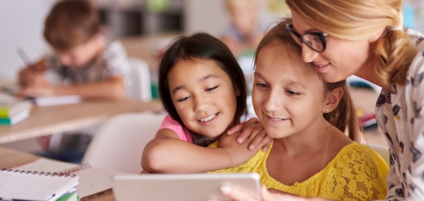 Eine Lehrerin zeigt zwei jungen Schülerinnen etwas auf dem Tablet. Sie sitzen in einem Klassenzimmer.