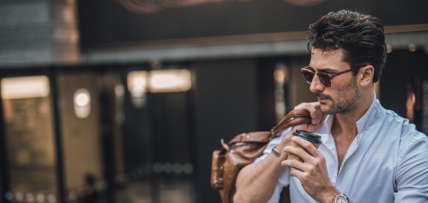 Junger Mann mit Sonnenbrille trinkt einen Kaffee während er seine Reisetasche über eine Schulter sowie eine Luxusuhr trägt. 