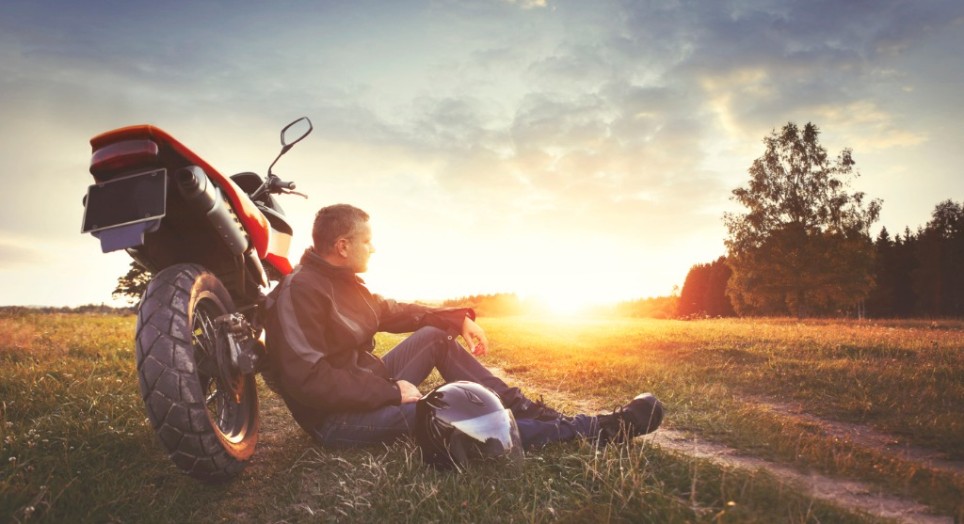 Ein Mann sitzt lehnend an seinem Motorrad auf einem Feld in der Sonne Ein Mann sitzt lehnend an seinem Motorrad auf einem Feld in der Sonne