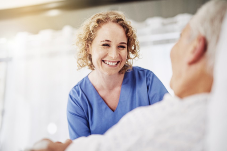 Eine Pflegerin schaut lächelnd einen im Bett liegenden Patienten an. 