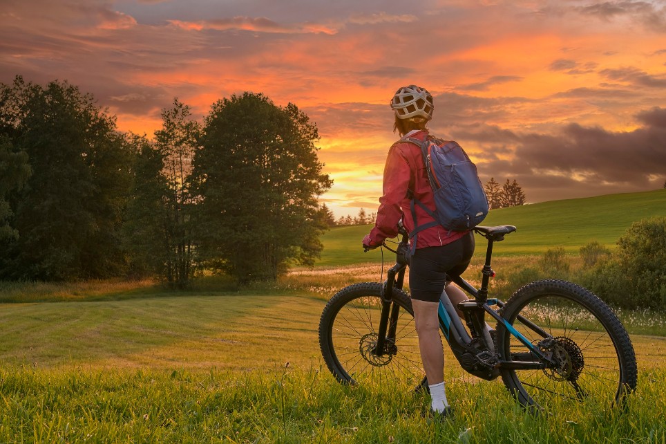 Eine Frau steht mit ihrem Mountainbike auf einem Feld und schaut in den Sonnenuntergang.