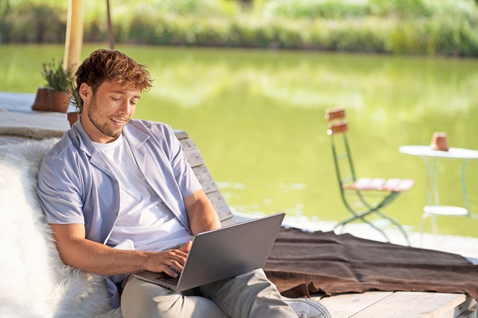 Junger Mann mit T-Shirt und Sommerhemd sitzt an einem See und schaut in ein Laptop