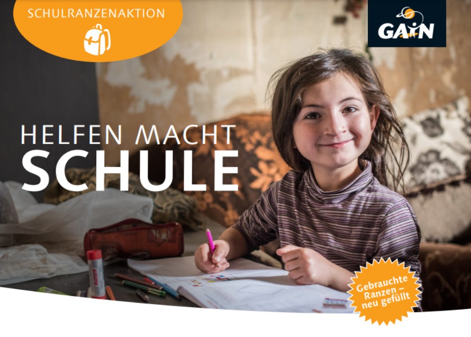 Schulranzenaktion Ein kleines Mädchen mit einem Heft und einem Stift in der Hand lächelt in die Kamera.