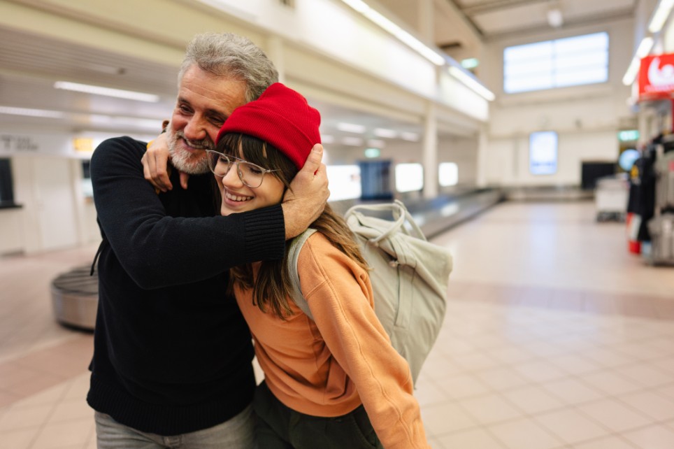 Vater mit grauem Bart und grauen Haaren begrüßt seine junge Tochter im Flughafen Foto einer jungen Frau und ihres älteren Vaters, die sich nach langer Zeit am Flughafen treffen und nach pandemiebedingten Trennungen und Isolation wieder zusammenkommen.