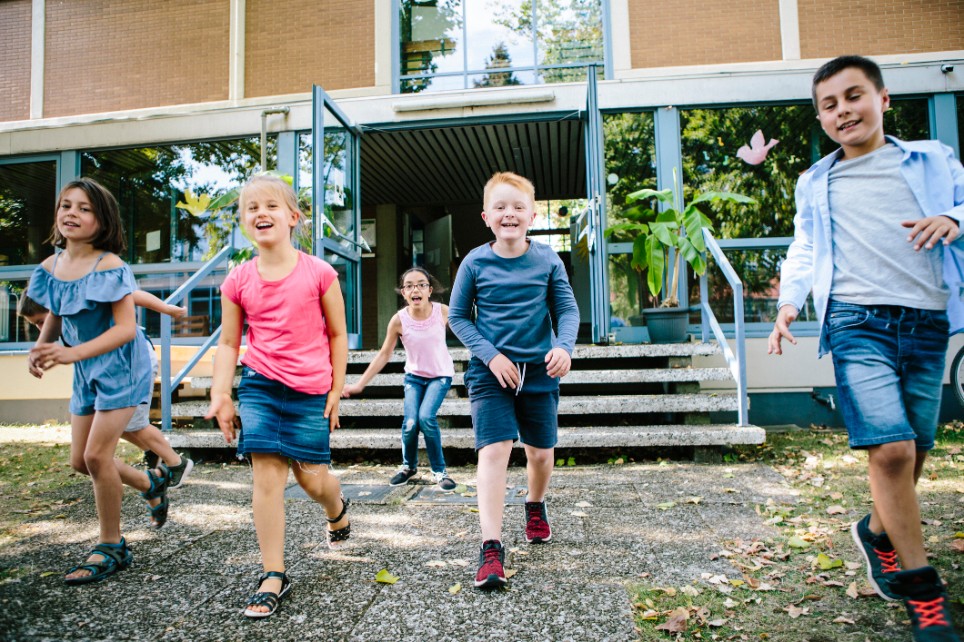 Fünf Kinder laufen lachend in die Kamera aus einen Schulgebäude raus 