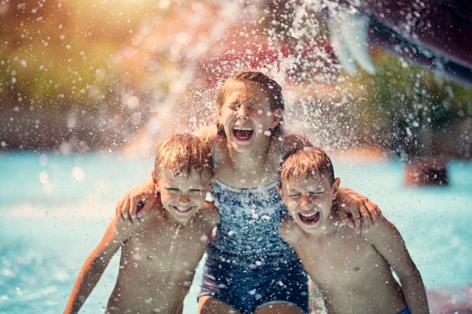 Drei Kinder stehen unter einem Wasserfall im Schwimmbad