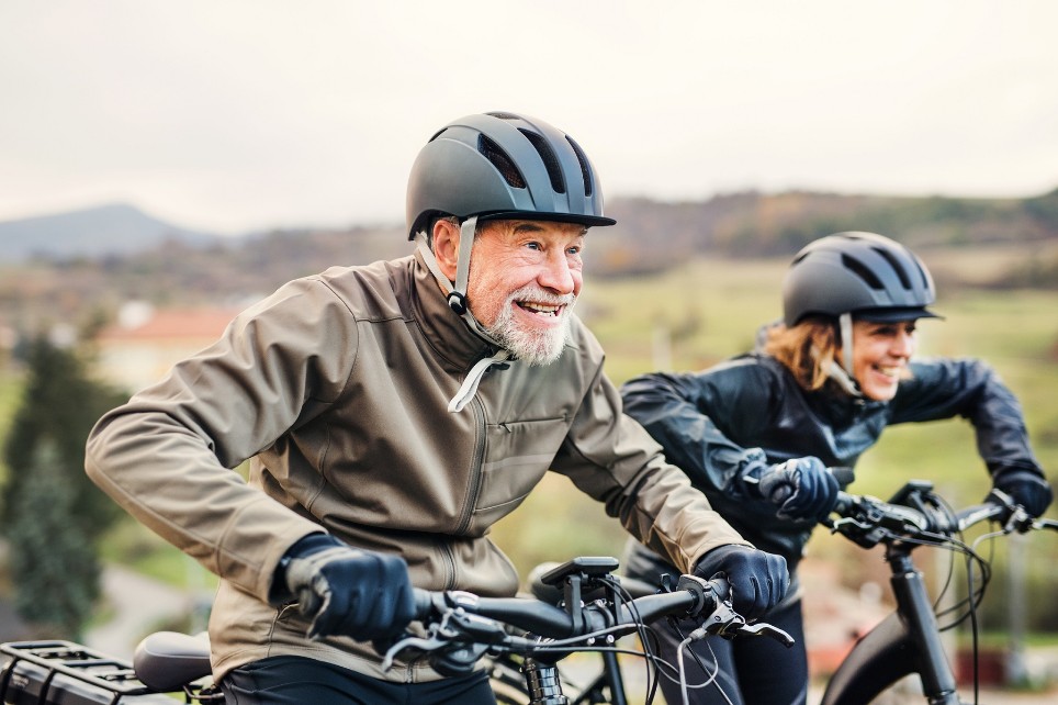 Eine älterer Mann und eine ältere Frau, beide mit Helm, sitzen startbereit auf ihren Fahrrädern. 