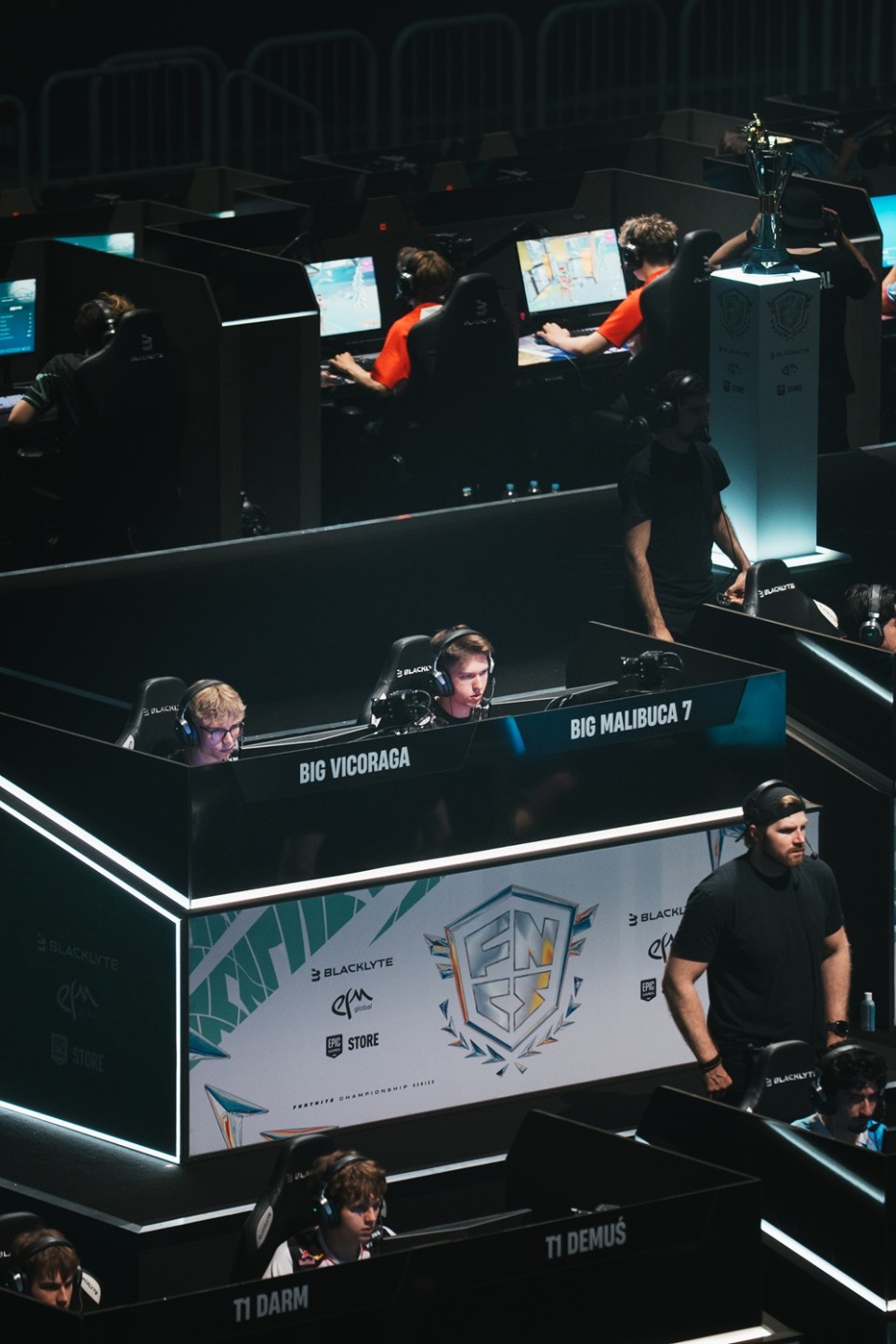 E-Sport-Spieler treten an mehreren Gaming-Stationen bei einem Fortnite-Turnier gegeneinander an; im Hintergrund steht der Siegerpokal.