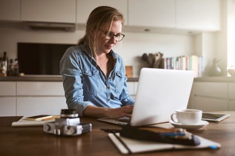 Eine Frau arbeitet mit ihrem Laptop und einigen Unterlagen am Esstisch in der Küche