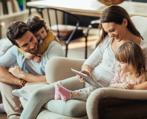 Eine junge Familie, bestehend aus 4 Personen, sitzt zusammen auf dem Sofa. Mutter und Tochter schauen in ein Tablet, Vater und Sohn schauen lächelnd zu ihnen rüber.