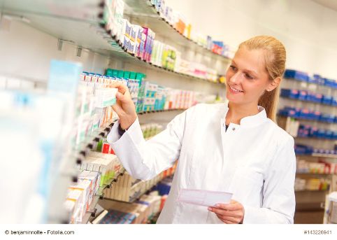 Eine junge Apothekerin hält ein Rezept in der Hand und greift gleichzeitig nach einem Medikament im Regal