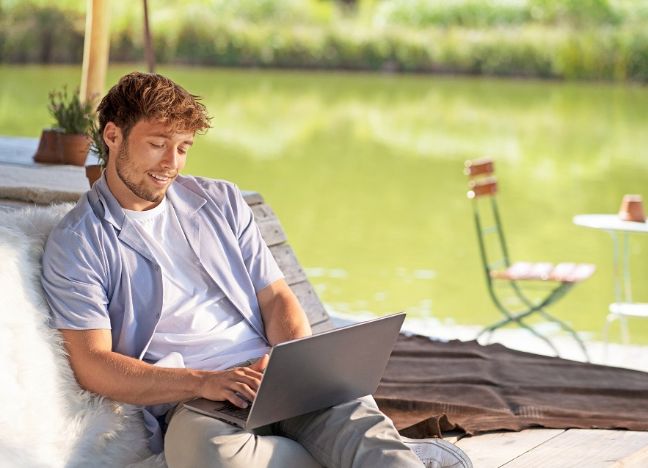Junger Mann mit T-Shirt und Sommerhemd sitzt an einem See und schaut in ein Laptop