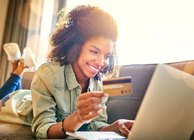 Eine junge Frau liegt auf dem Sofa und tippt lächelnd die Daten ihrer Kreditkarte in ihren Laptop ein.