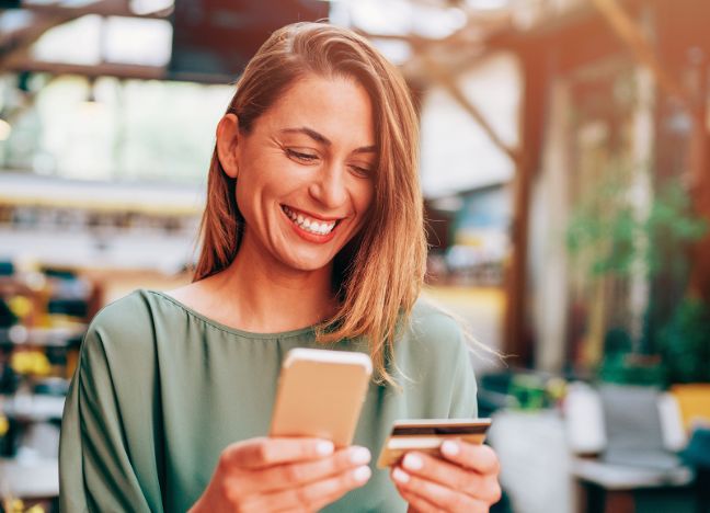 Eine junge Frau tippt lächelnd die Daten ihrer Kreditkarte in ihr Smartphone ein.